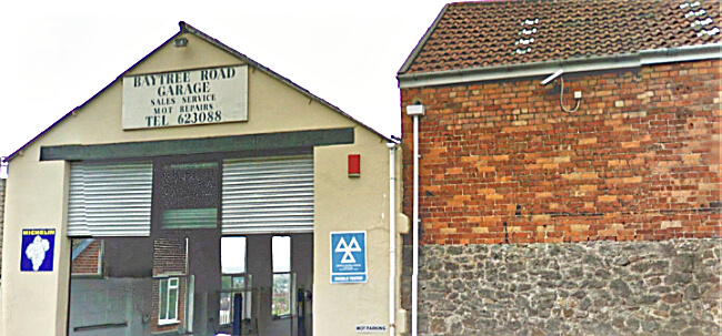 The gable end of Baytree Road Garage showing the hand-painted sign reading Baytree Road Garage, Sales Service, MOT Repairs, Tel 623088, with the roller shutter and blue MOT triangle beside the archway entrance.